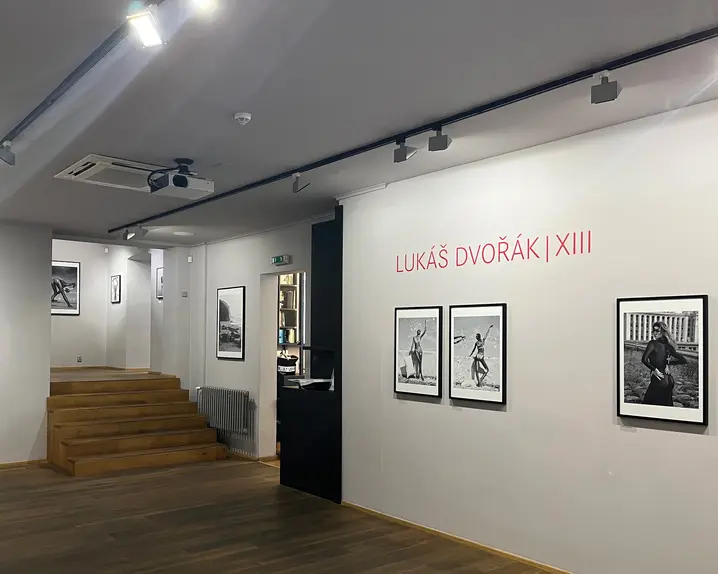 Gallery of black and white photographs by Lukáš Dvořák, showing women in various places. A wooden staircase leads to another room with photographs.