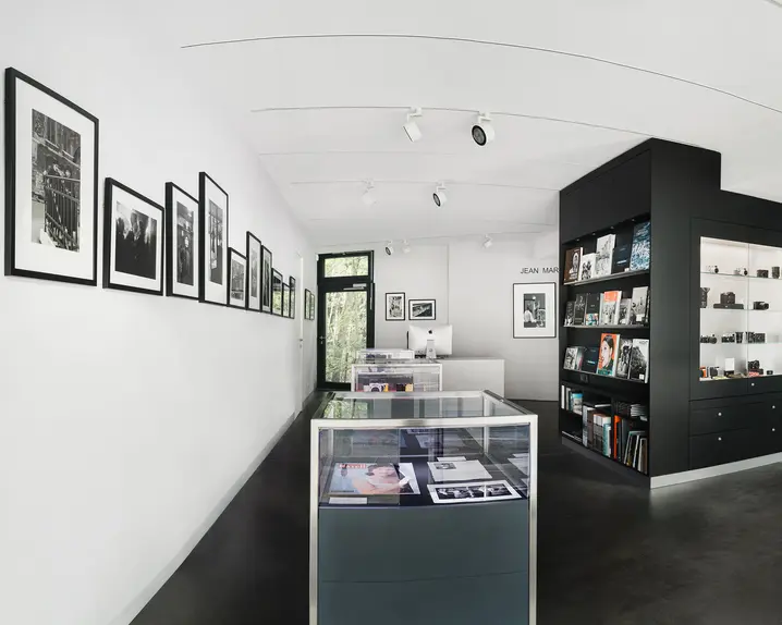 Bright exhibition room with black and white photographs, bookshelves and glass cabinets displaying cameras.