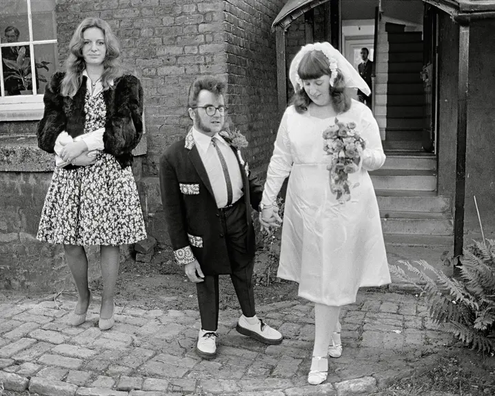 A wedding couple and a woman in a dress stand in front of a building.