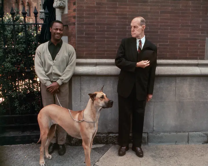 Two men, one with a dog on a lead, stand in front of a brick wall.