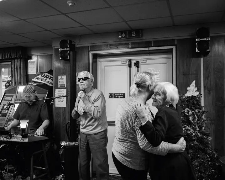 A man sings into a microphone while two older women dance and a man plays the keyboard in the background.