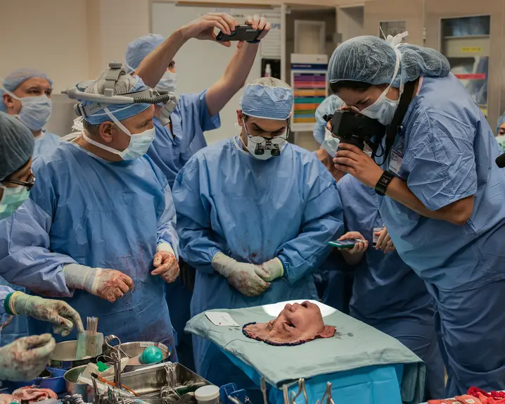 A group of people in surgical gowns look at a mask representing a face, and some take pictures of it.