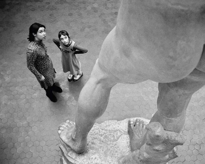 Two people stand in front of a large statue and look upwards.