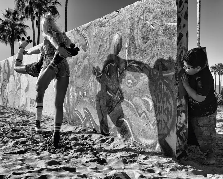A boy looks behind a graffiti-painted wall at two women on roller skates warming up.