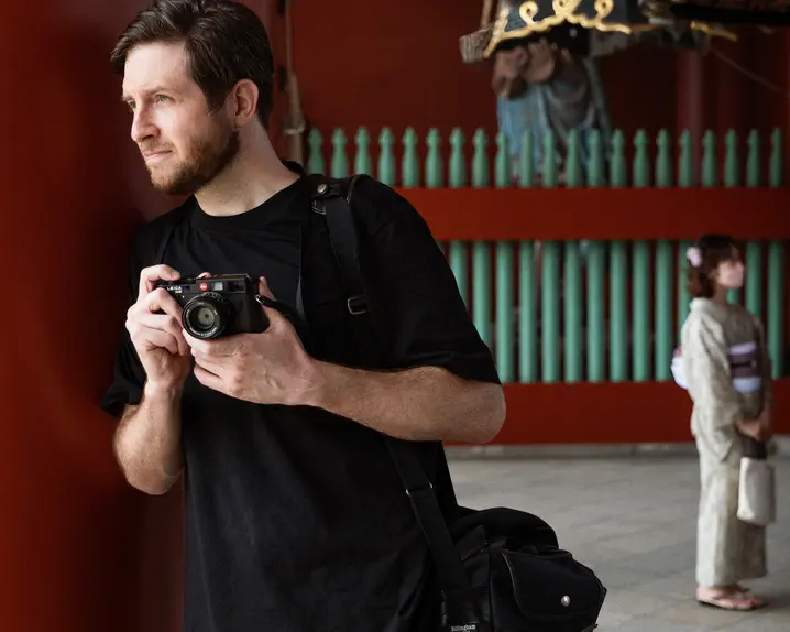 A photographer holds a camera while standing near traditional architecture, with two people in kimonos in the background.