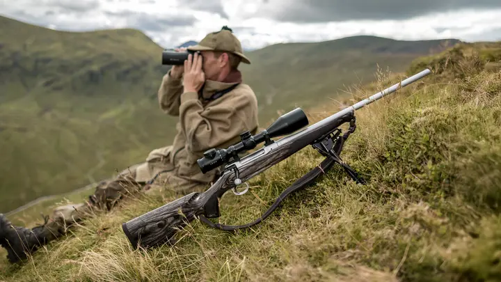 A man sitting on a hill with binoculars. In front of him is the Leica Magnus riflescope mounted on a weapon