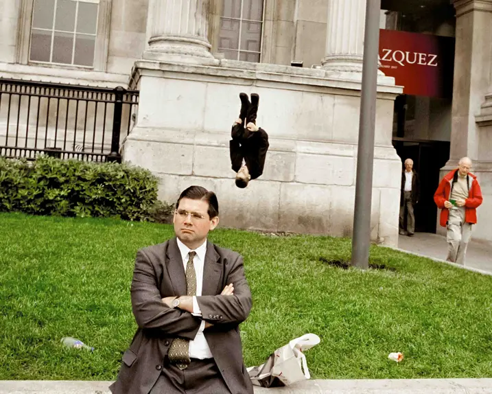Man in a suit sits on a wall behind him a man in a suit does a somersault