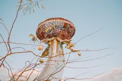 Metallic sculpture with colourful details in front of a blue sky and branches.