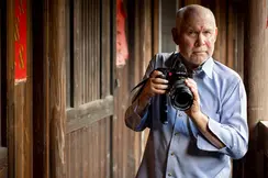 Steve McCurry holds a Leica camera and stands in front of a wooden house.