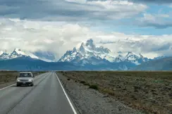 A car is driving on a road with snow-covered mountains in the background.