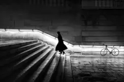 A woman in a long coat is walking up a staircase at night. The banister is illuminated.