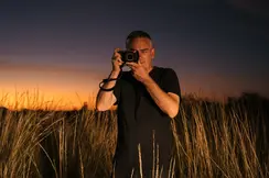 Person in a field with tall grass taking photos with a camera at sunset.