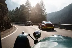 Vintage cars drive along a winding mountain road surrounded by trees and rocks.
