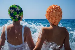 Two people in swimsuits and colorful bathing caps sitting in splashing water in the sea.