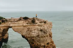 A person is standing alone on a rocky cliff jutting out into the ocean, and it is raining.