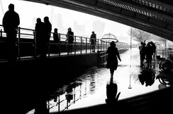 People walking under a bridge in the rain, silhouettes reflected in puddles.