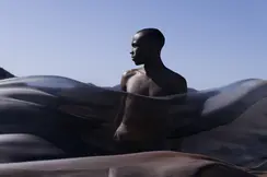 Man behind a transparent cloth looks to the side and light blue sky in the background.