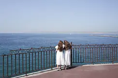 Two girls in white dresses stand at a railing and look to the sea.