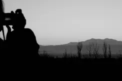 Black-and-white photograph of a person taking a picture of a lake and mountains in the background.