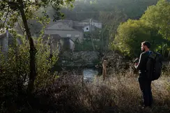 A man with a rucksack stands in the countryside and looks out over a small settlement by the river