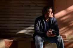 A man sits on a step with a camera in his hands, his face in the sunlight, in front of a closed shutter.