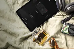 A camera, tablet, headphones, sunglasses, perfume and wallet laying on a bedcover.