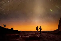 A couple walks in the desert under a starry sky toward a orange horizon.