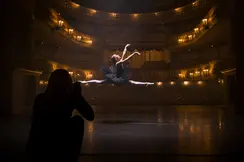 A person in the shade takes a picture of a ballerina jumping in an illuminated theatre.