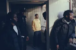Five men in a shady hallway looking outside, the one in the middle wearing a yellow jacket.