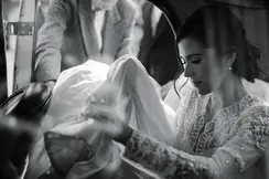 A bride sitting in a car holding her veil and a man standing next to the open car door.