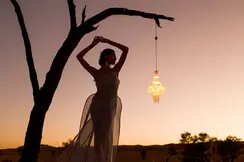 Woman posing under a tree at sunset next to a hanging lantern.