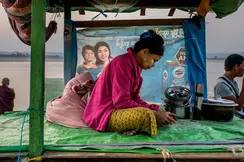 Woman sits on plastic tarp looking at a phone, in background are other people and a river.