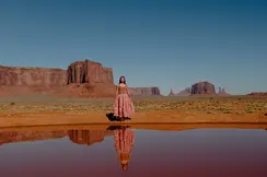 Woman standing in a desert landscape with red rock formations in the background, the image is reflected in the water in front of her