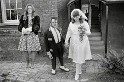 A wedding couple and a woman in a dress stand in front of a building.