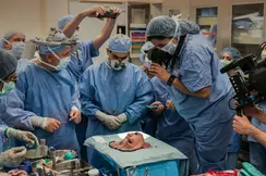 A group of people in surgical gowns look at a mask representing a face, and some take pictures of it.