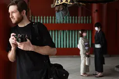 A photographer holds a camera while standing near traditional architecture, with two people in kimonos in the background.