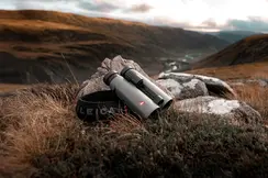 The Leica Noctivid leans against a medium-sized stone, while a picturesque, almost stormy landscape stretches out in the background.