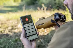 A mobile phone held by a hunter, opened with Leica Ballistics App and Leica Geovid Pro AB+ in the foreground, a meadow can be seen in the background.