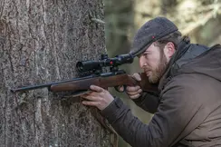 The Leica Fortis 6 1.8-12x42i is in focus while a hunter leans against a tree with his weapon and looks through the scope