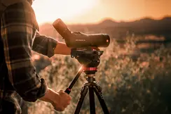 The Leica APO Televid is in the foreground while being operated by a man in a lumberjack shirt. In the background is a picturesque landscape at sunset