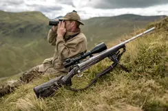 A man sitting on a hill with binoculars. In front of him is the Leica Magnus riflescope mounted on a weapon