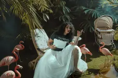 Woman with white tulle skirt and cigarette in her hand on a lounger with flamingos standing around her