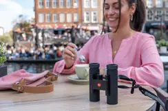 Woman is sitting in a cafe with her Leica Noctivid Compact next to her