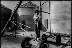 Young girl standing beside farm equipment and grain silos on a family farm in Arkansas, captured in black and white. A documentary photograph reflecting rural life, agricultural heritage, and generational connection.