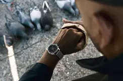 Jason Roman checks his Leica Watch while feeding pigeons in an urban public space, capturing a candid moment of everyday city life and human interaction with wildlife.