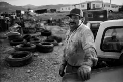 A man in a worn work shirt leans against a pickup truck in a rural scrapyard in Montana. Old tires, machinery, and trailers fill the foreground, with the Rocky Mountains in the distance.