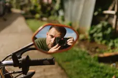 Photographer Philipp Gladsome reflected in a motorcycle mirror while taking a picture with a Leica M camera, surrounded by greenery in warm natural light.