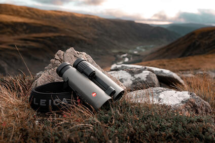 Leica Noctivid rest on a rugged rock, overlooking a serene mountain valley at dusk.