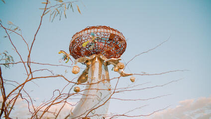 Metallic sculpture with colourful details in front of a blue sky and branches.