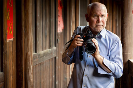 Steve McCurry holds a Leica camera and stands in front of a wooden house.